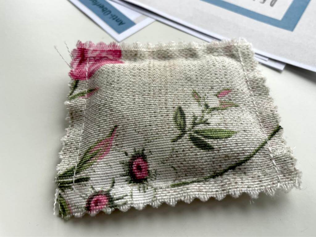 Lavendelduftkissen: Ein kleines genähtes Stoffkissen mit grauem Grundton mit rosa Blumen darauf. Im Hintergrund zwei Blätter Papier.